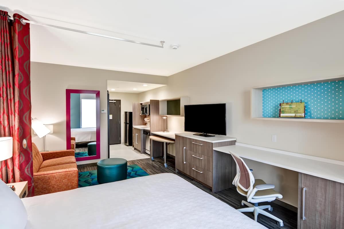 A hotel room with a bed, a desk, a TV, a mirror, and a kitchenette.