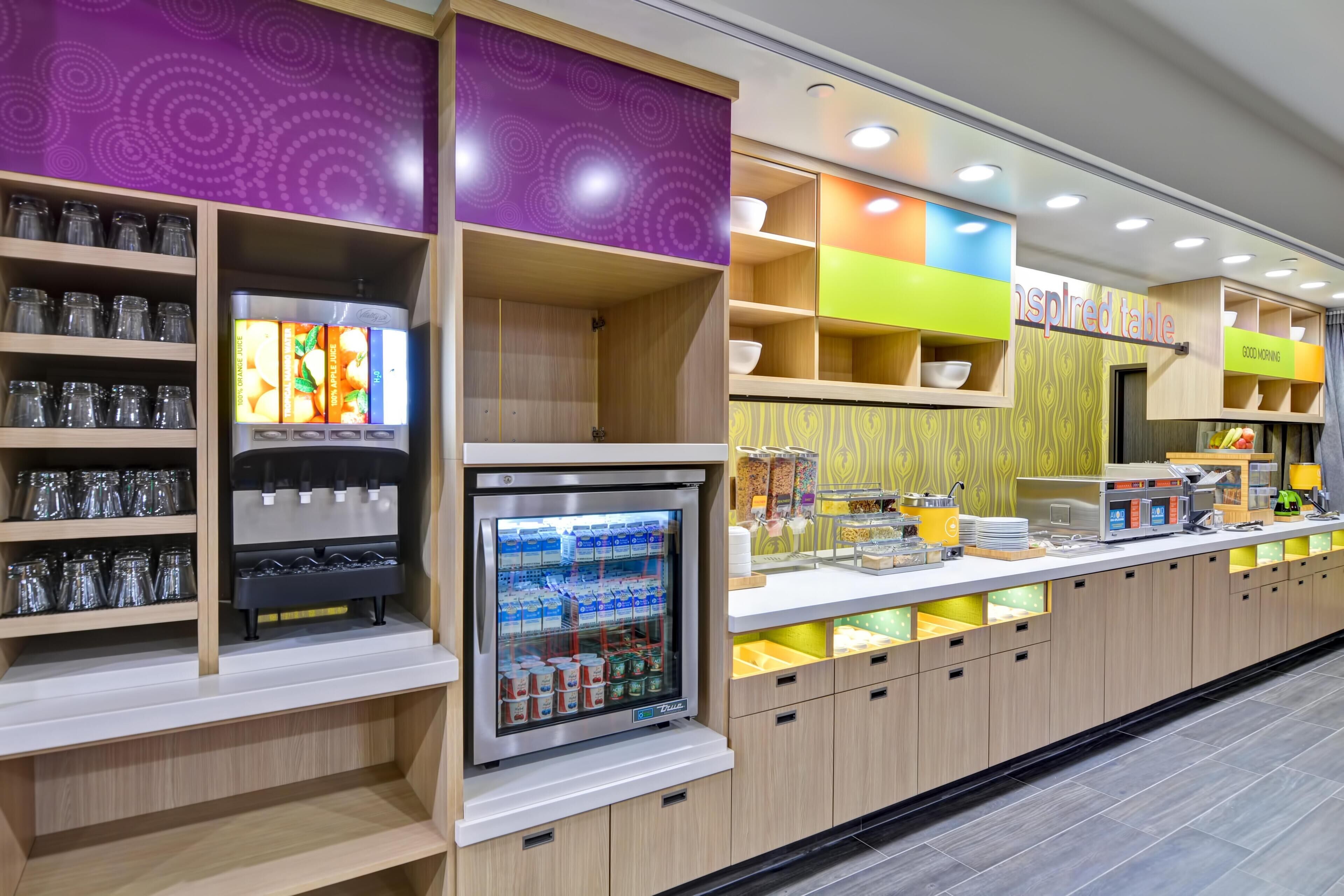 A self-service breakfast area with beverage dispensers, refrigerated display, and various food items.