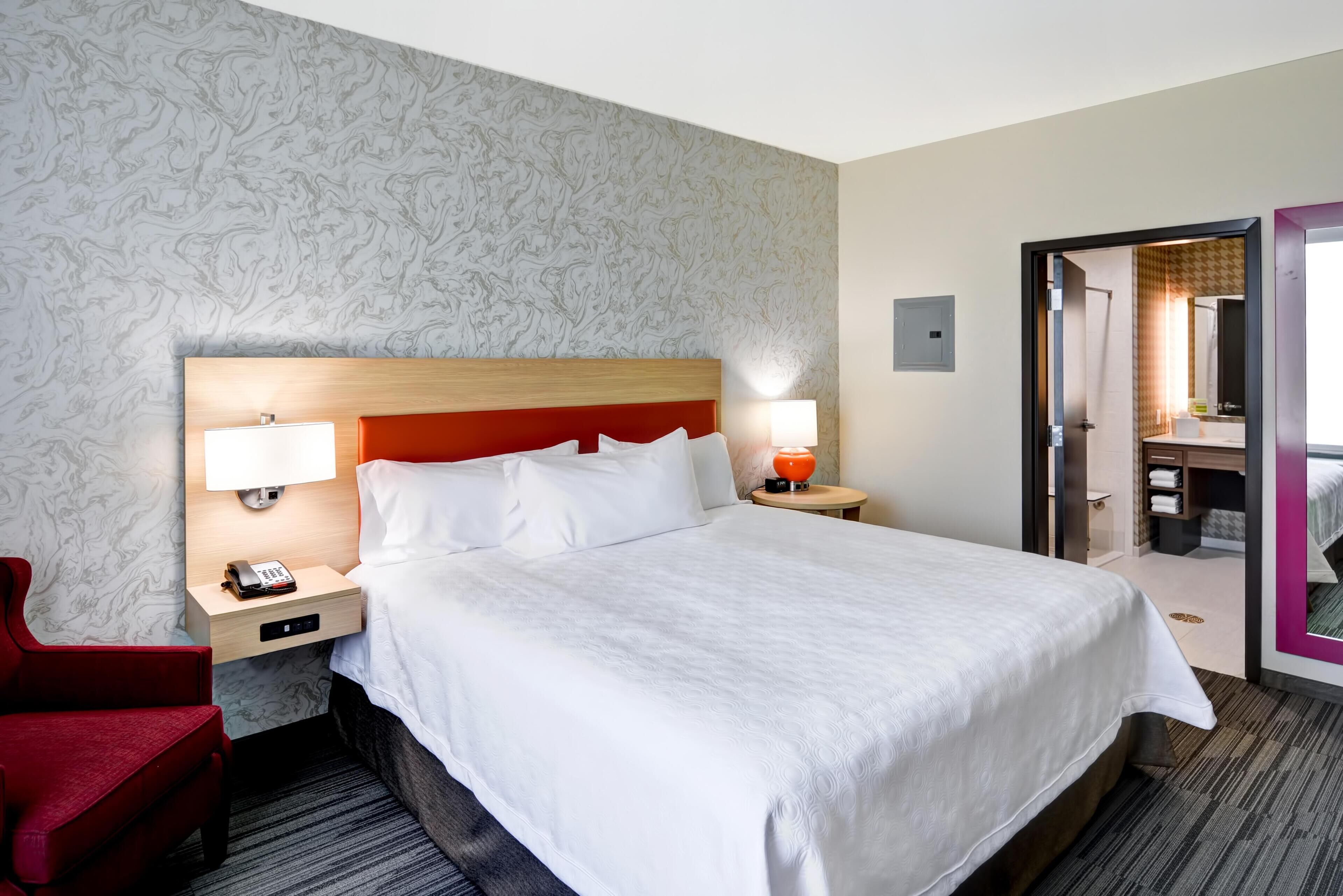 A hotel room with a large bed, a red chair, a nightstand with a phone, and a mirror.