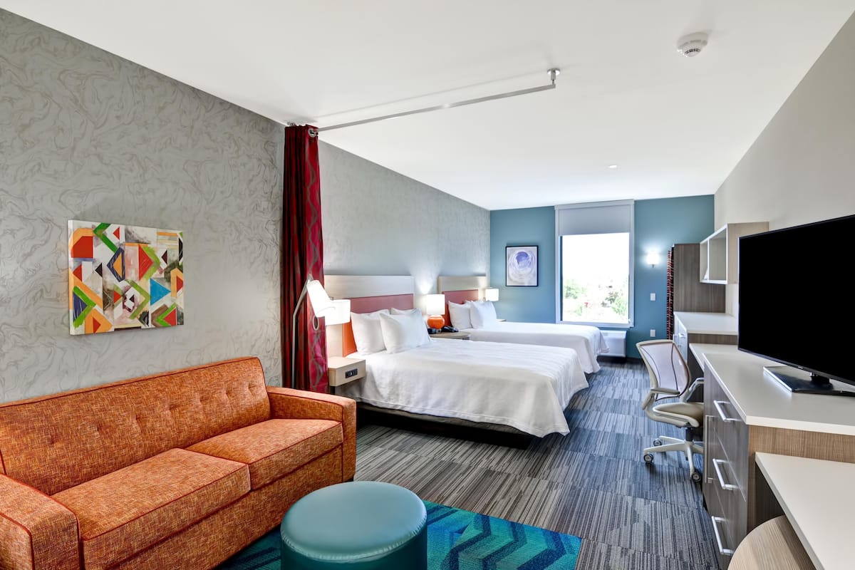 A hotel room with a sofa, two beds, a desk, a TV, and artwork on the walls.