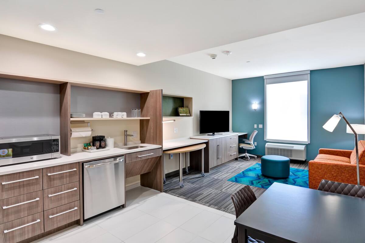 A modern kitchen with a microwave, sink, and dishwasher, a dining area with a table and chairs, and a TV in a room with blue walls and a large window.