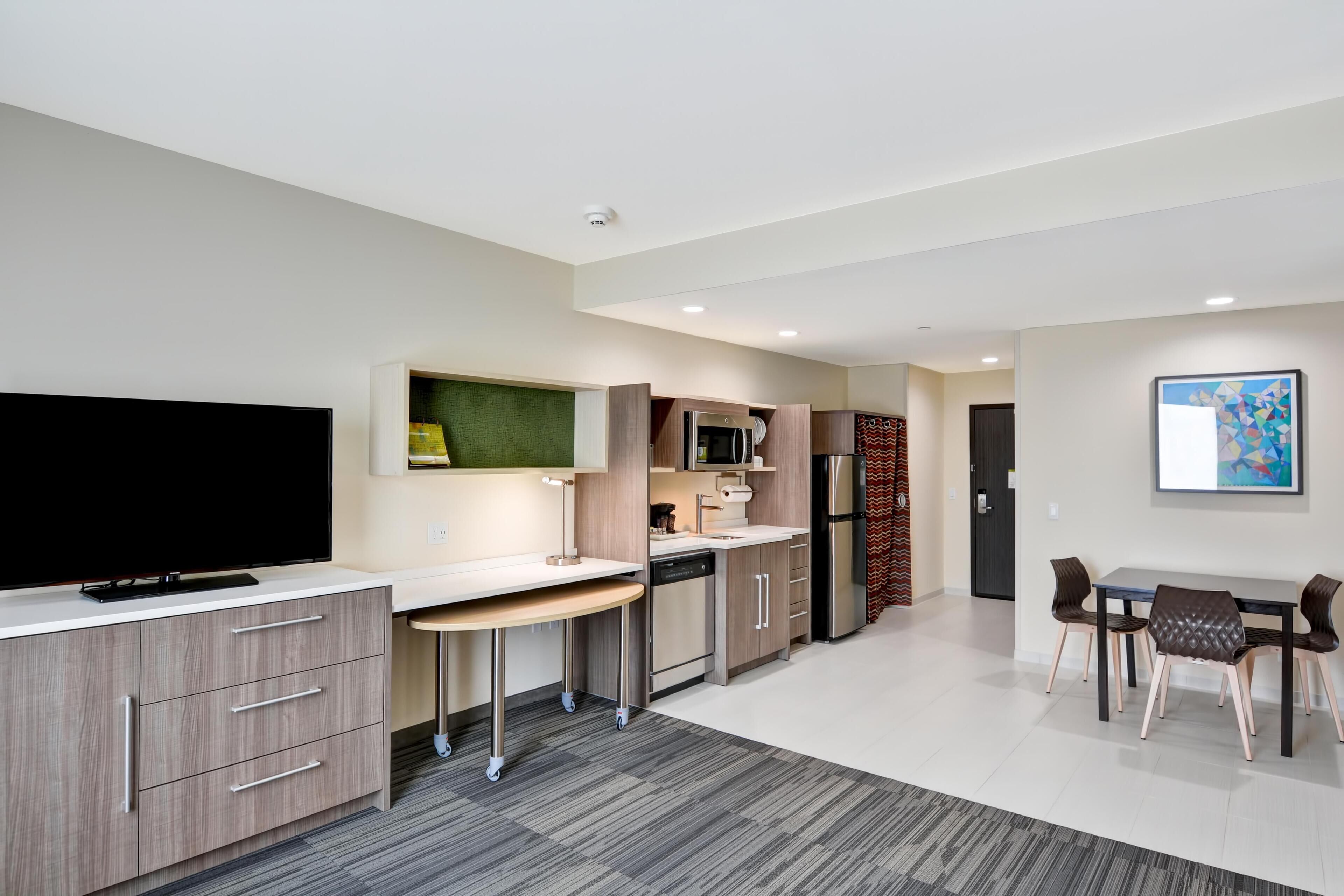 A modern kitchen with a flat-screen TV, a microwave, and a refrigerator. There is a dining area with a table and chairs.