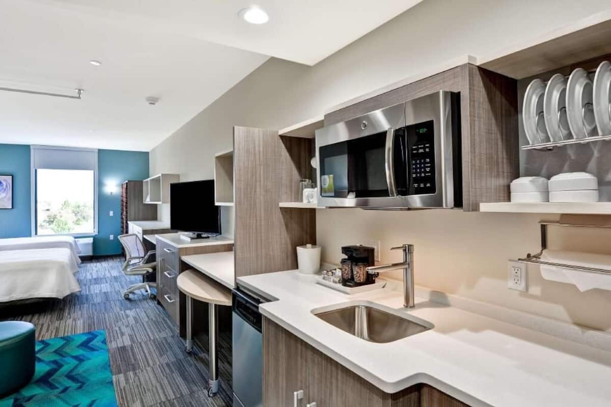 A modern hotel room with a kitchenette, a bed, a desk, and a view of the outdoors.