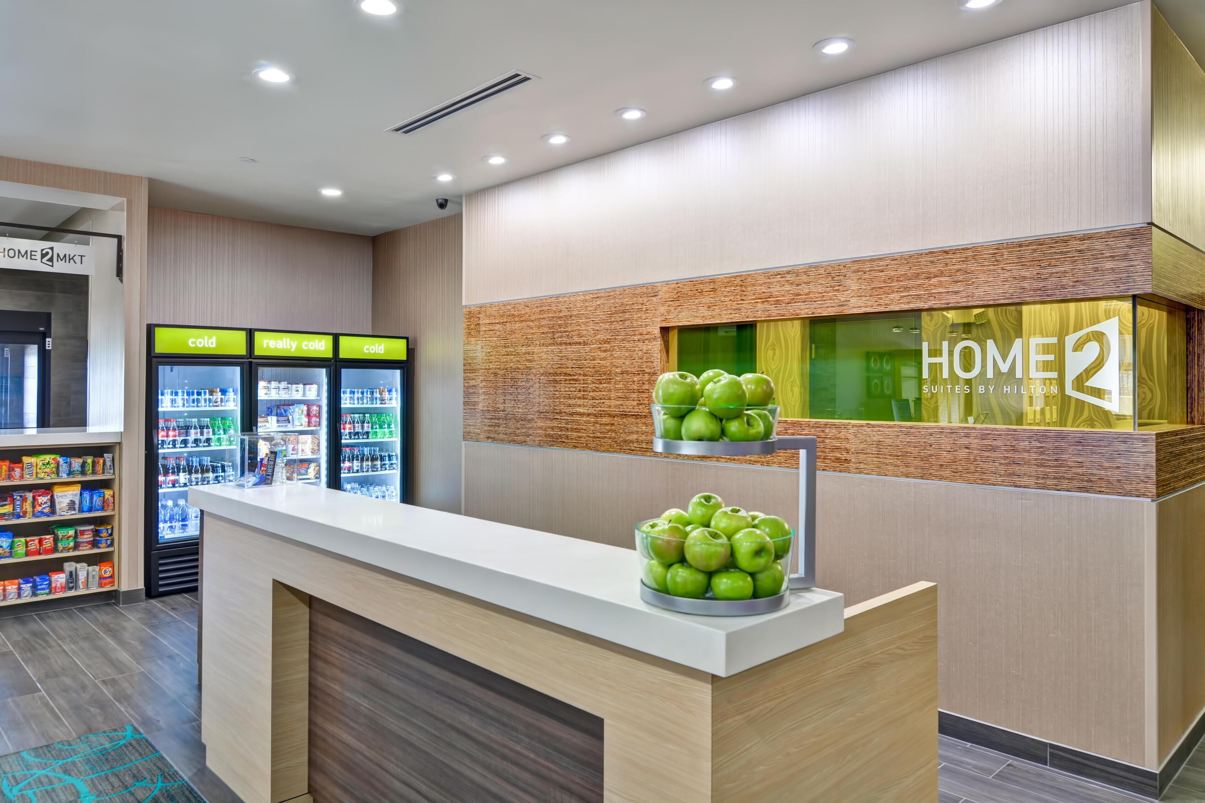 Zona de recepción de un hotel con mostrador, una exhibición de manzanas verdes y un cartel que dice 'HOME 2 SUITES BY HILTON'.