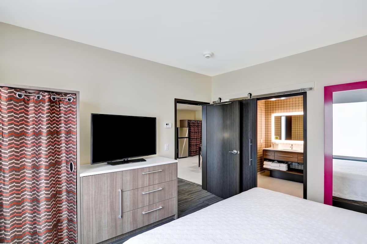 A bedroom with a bed, a TV on a dresser, a wardrobe, and a bathroom visible through an open door.