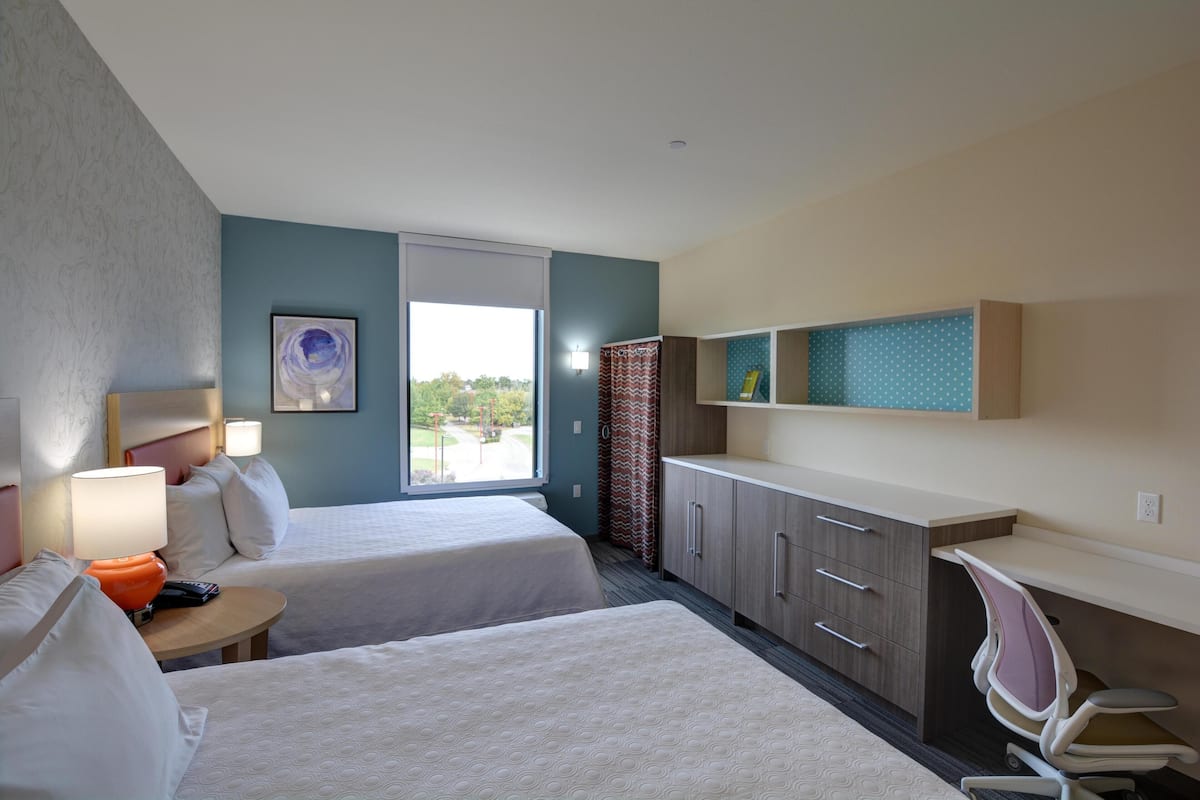 A hotel room with two beds, a desk, a chair, a lamp, a nightstand, and a window with a view.