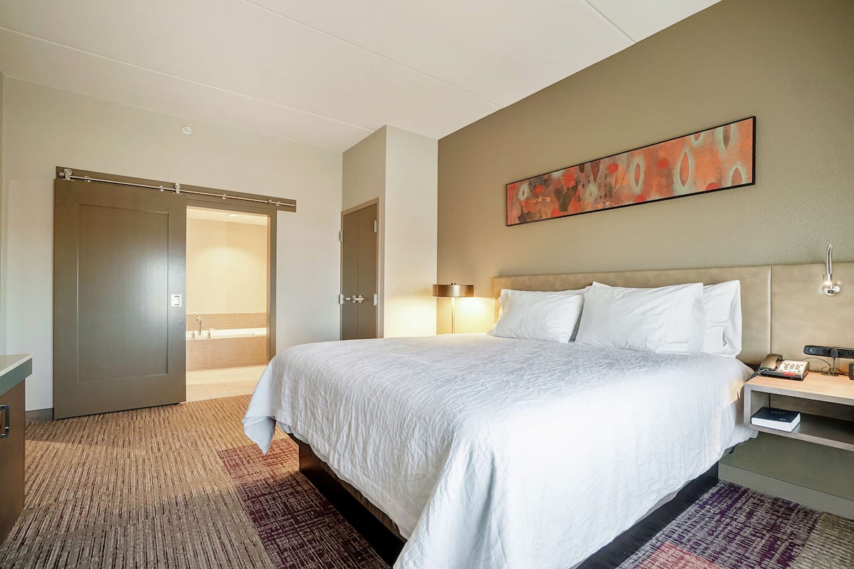 A hotel room with a large bed, a nightstand, a wall-mounted lamp, and a door leading to a bathroom.