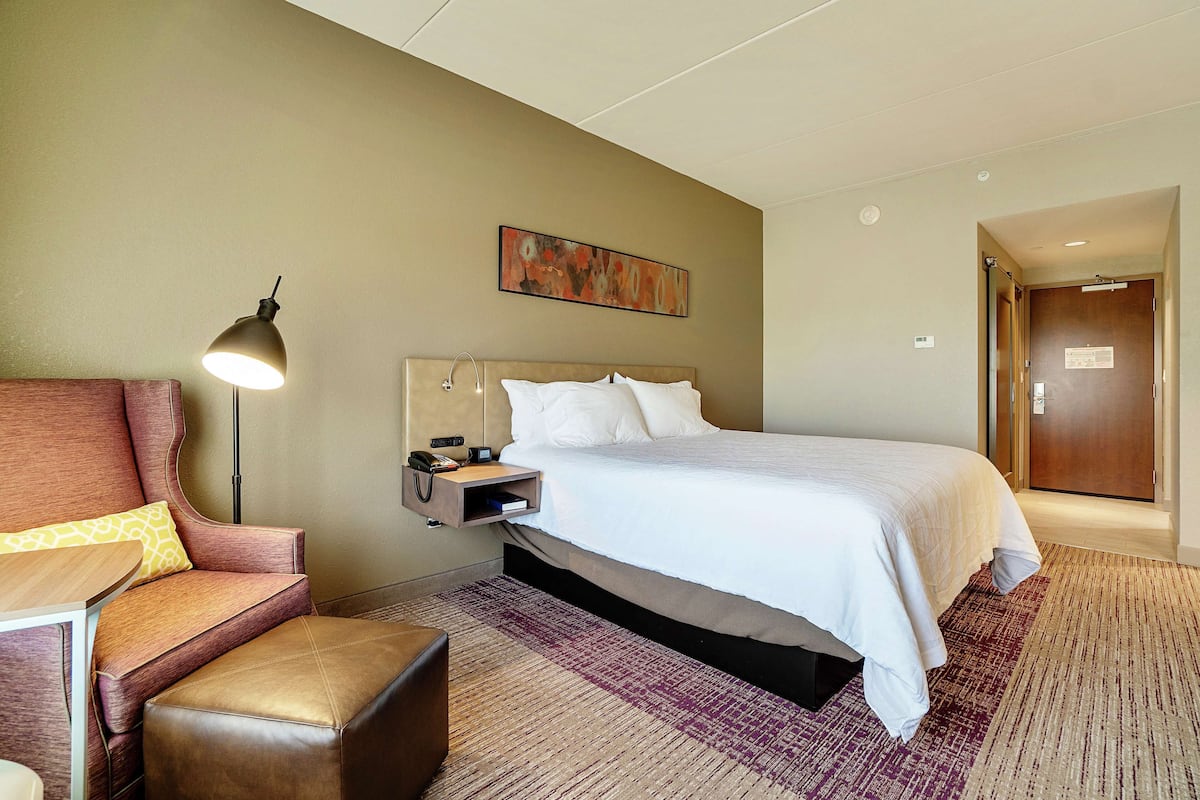 A hotel room with a bed, a desk with a lamp, a chair, and a small table.