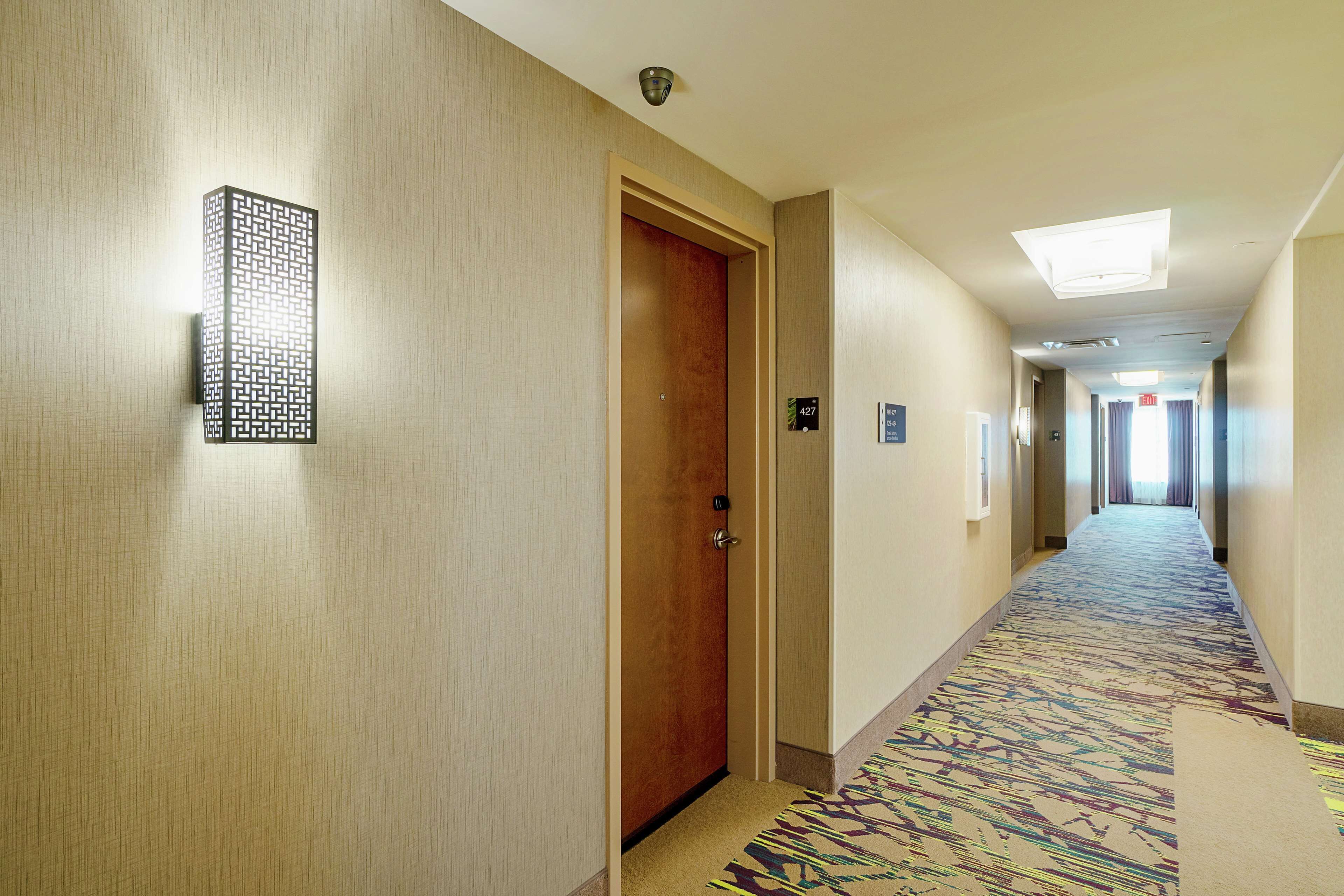 A hotel hallway with a patterned carpet, a wooden door, and a wall-mounted light fixture.