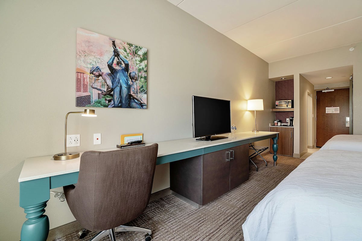 A hotel room with a bed, a desk with a computer, a chair, a TV, and a painting on the wall.