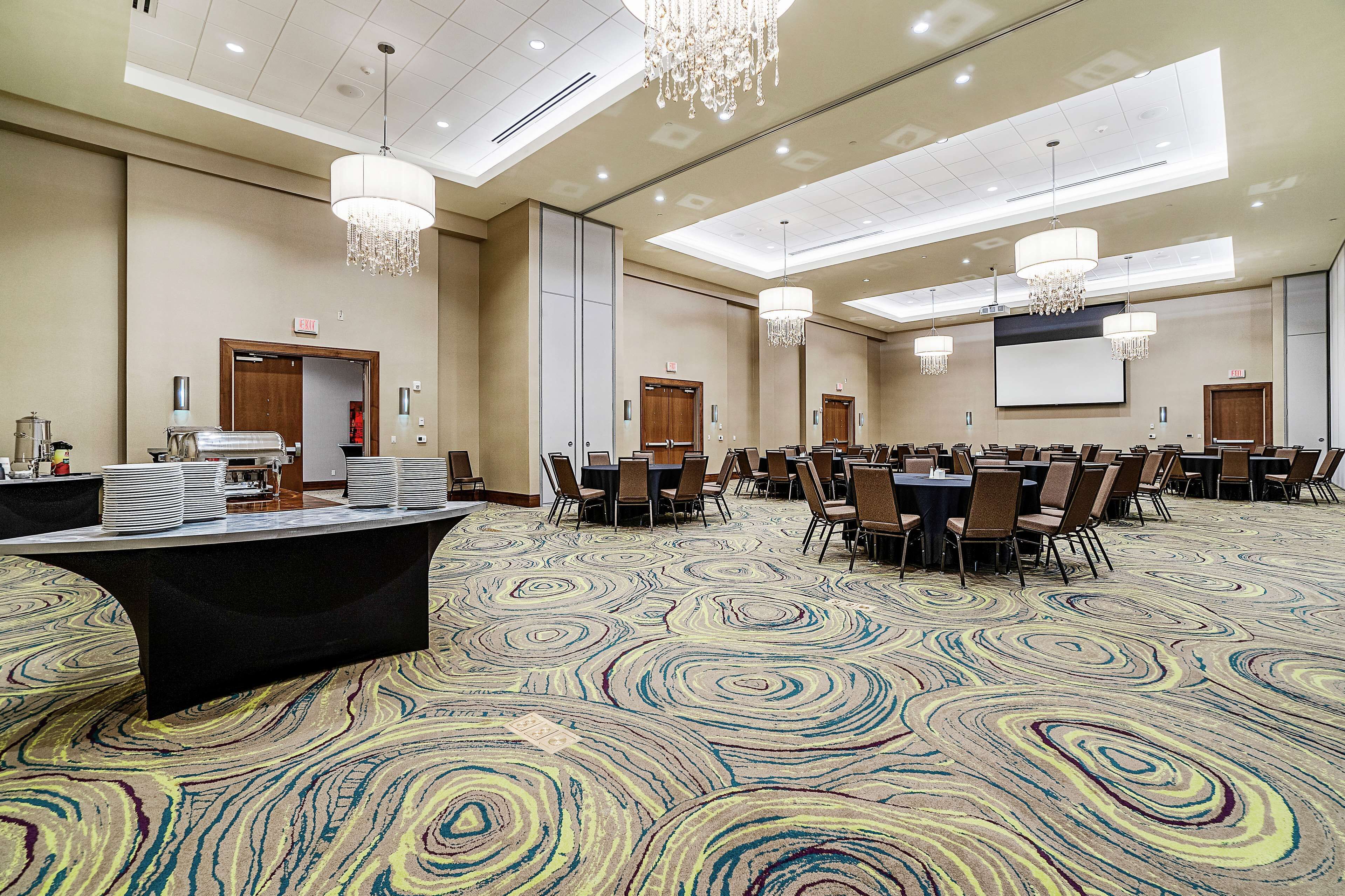 A conference room with a large carpeted floor, multiple tables with chairs, and chandeliers.