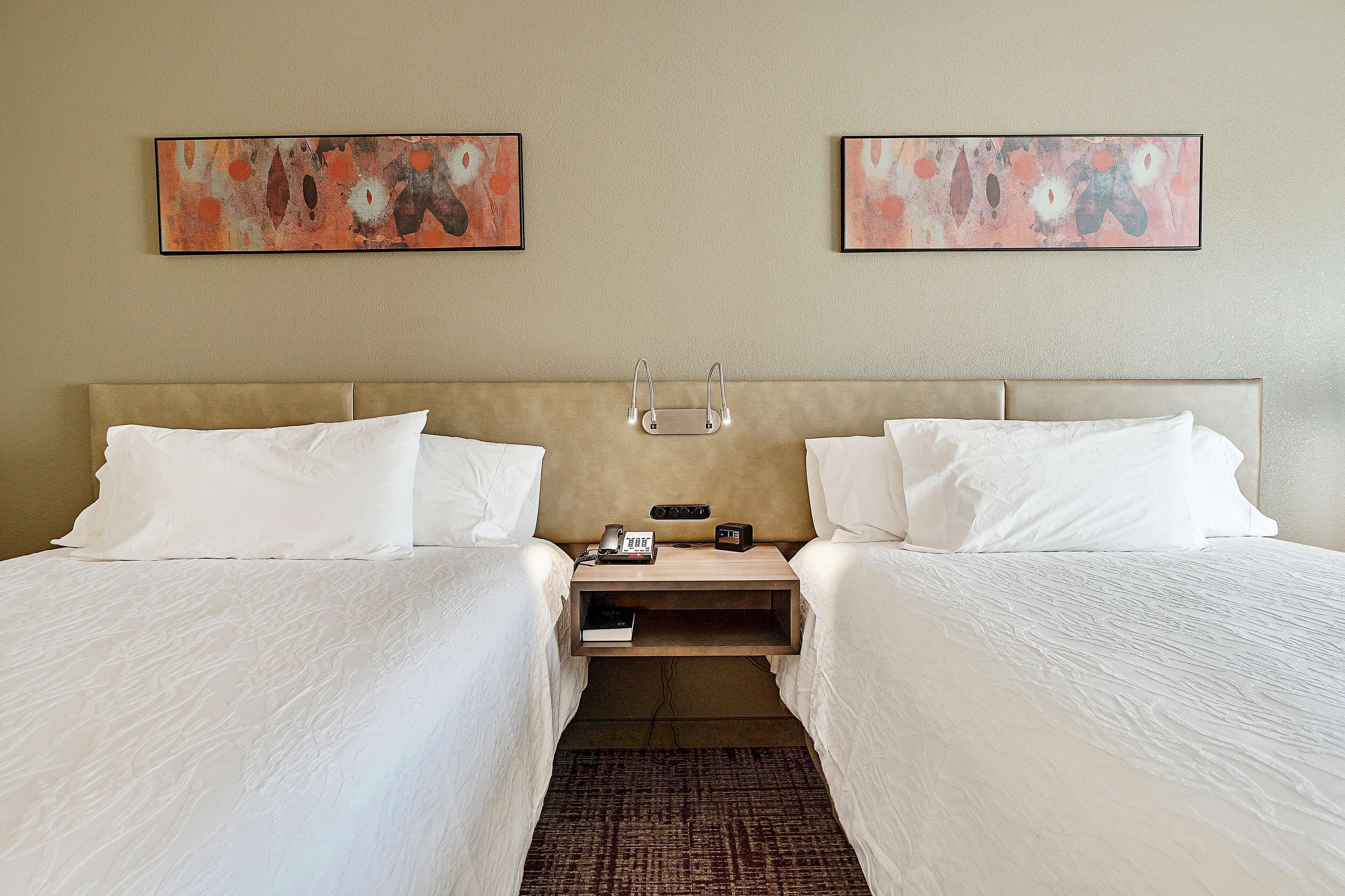 Une chambre d’hôtel avec deux lits, une table de chevet sur laquelle se trouvent un téléphone et un livre, et des tableaux abstraits accrochés au mur.
