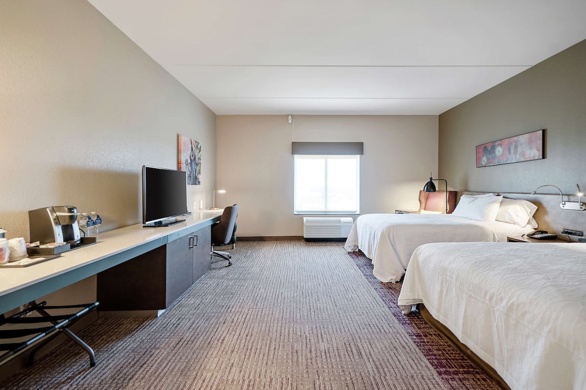 A hotel room with a desk, two beds, a TV, a window, and a painting on the wall.