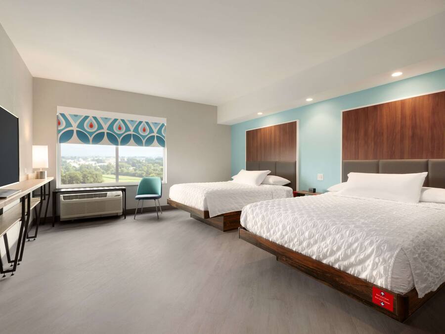 A modern hotel room with two beds, a desk, a chair, and a large window offering a city view.