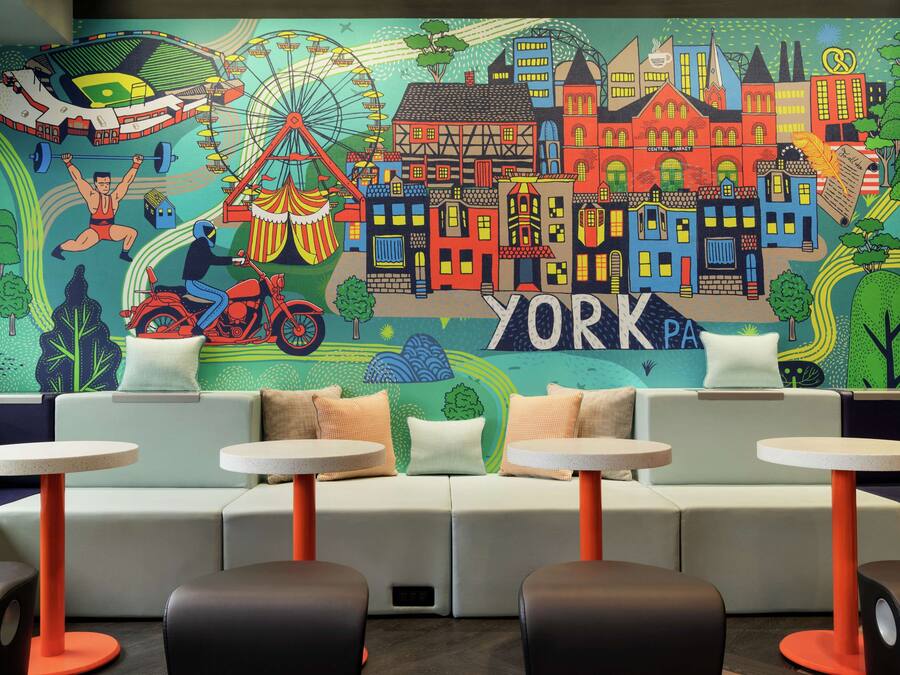 A modern lounge area with a colorful mural of York, featuring a Ferris wheel, buildings, and a cyclist. There are sofas and round tables with orange legs.