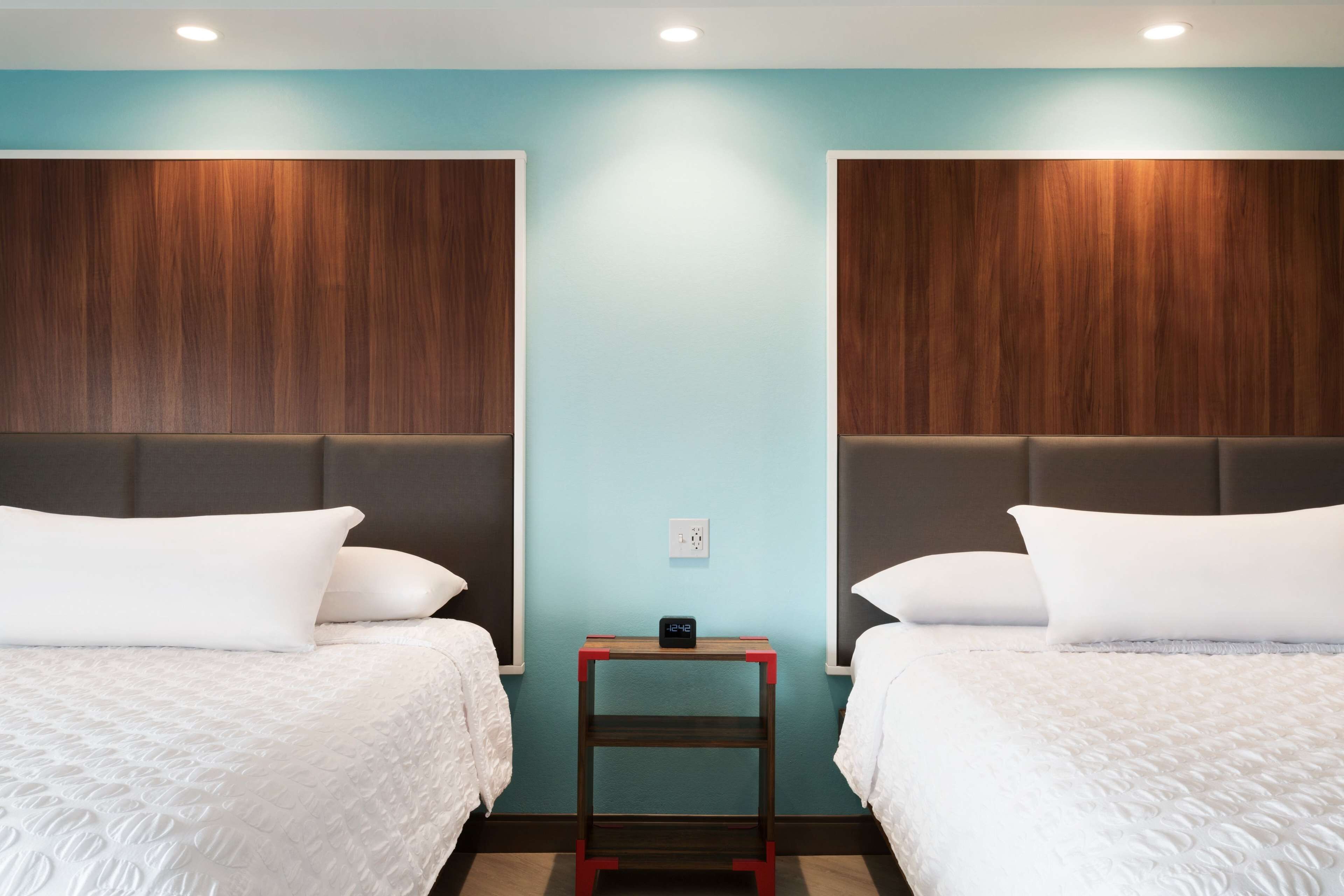 A hotel room with two beds, a nightstand with a clock, and a light blue wall.