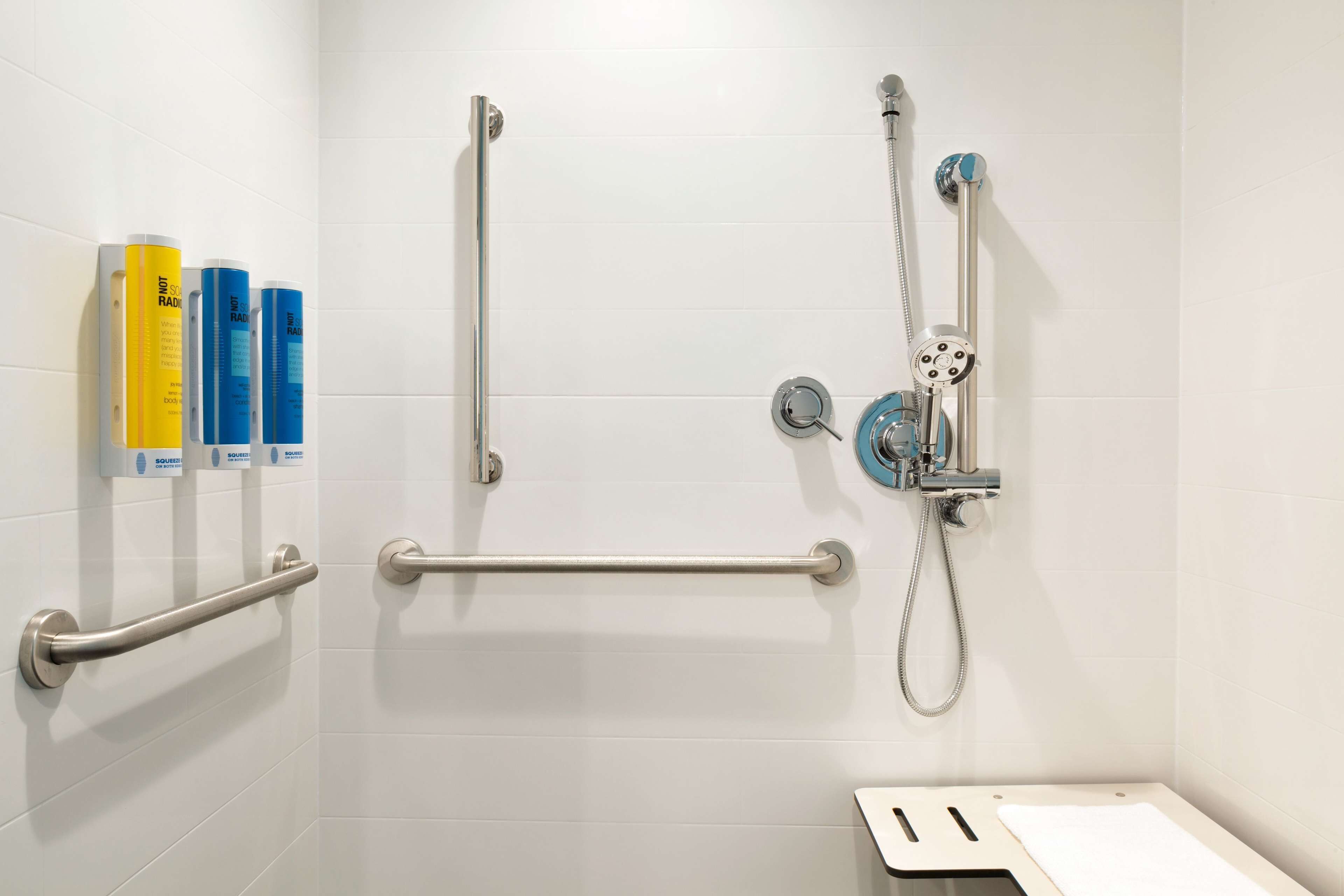 Une salle de bain avec des murs carrelés de carreaux blancs, une douche équipée d’un pommeau de douche à main et de deux barres d’appui.