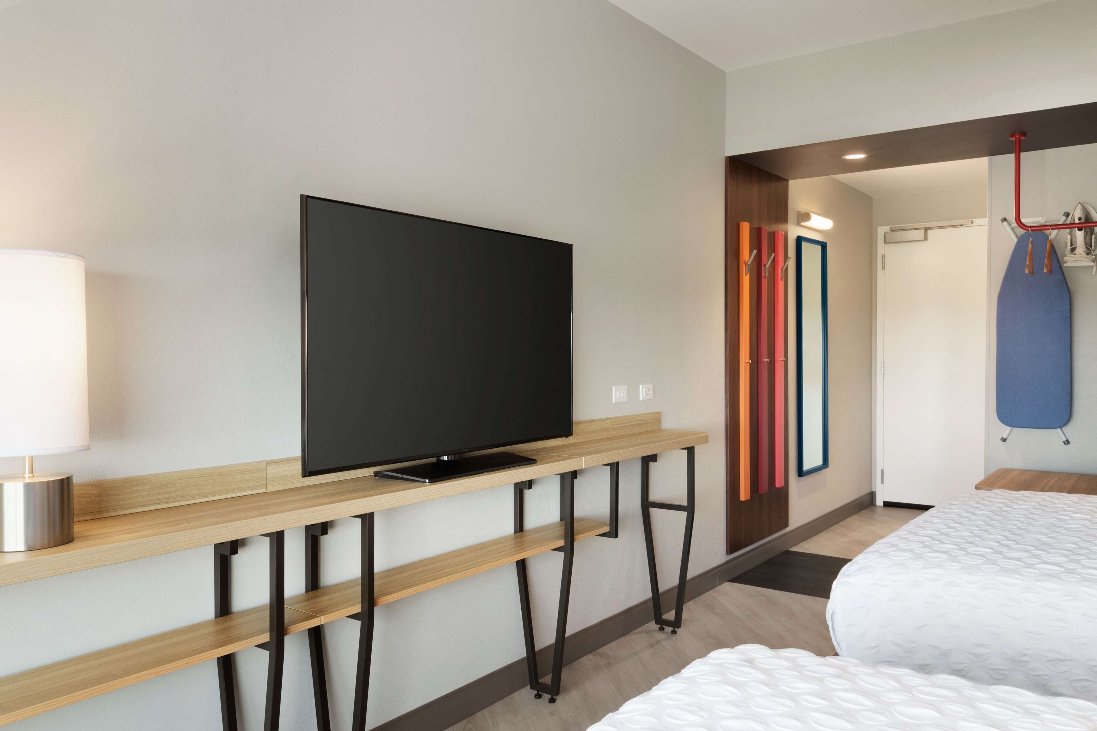 A modern hotel room with a flat-screen TV on a wooden console table, a bedside table, a bed with a white quilt, and a door with colorful vertical blinds.