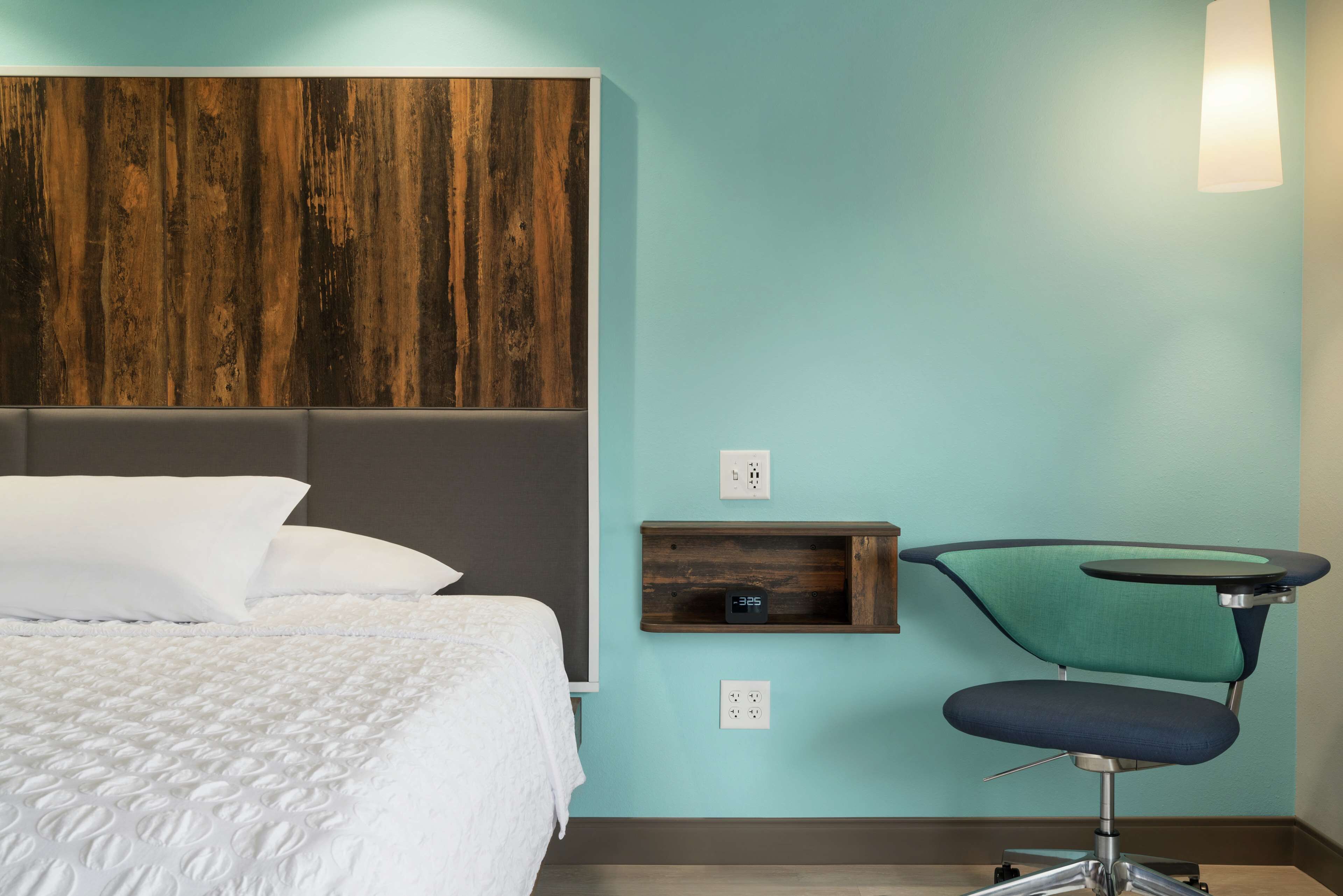 A bedroom with a bed, a wooden headboard, a teal wall, a small shelf, and a chair.