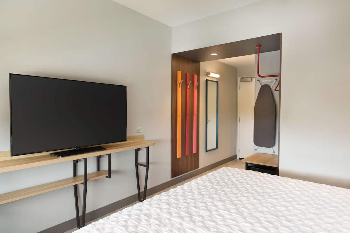 A modern hotel room with a flat-screen TV on a wooden console, a bed with a white quilt, a mirror, and a red ironing board.
