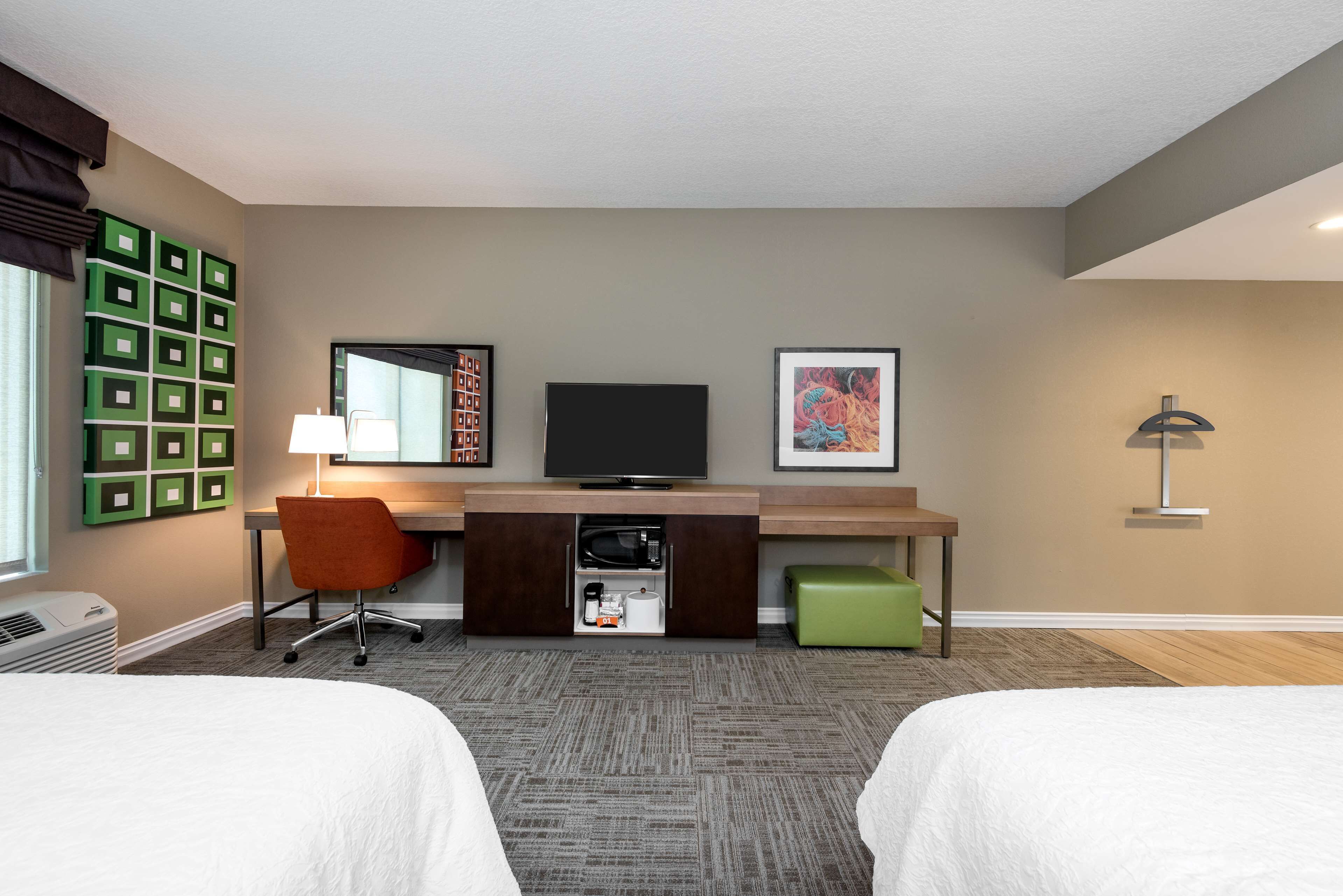 A hotel room with two beds, a desk with a television, a chair, a lamp, and a green ottoman.