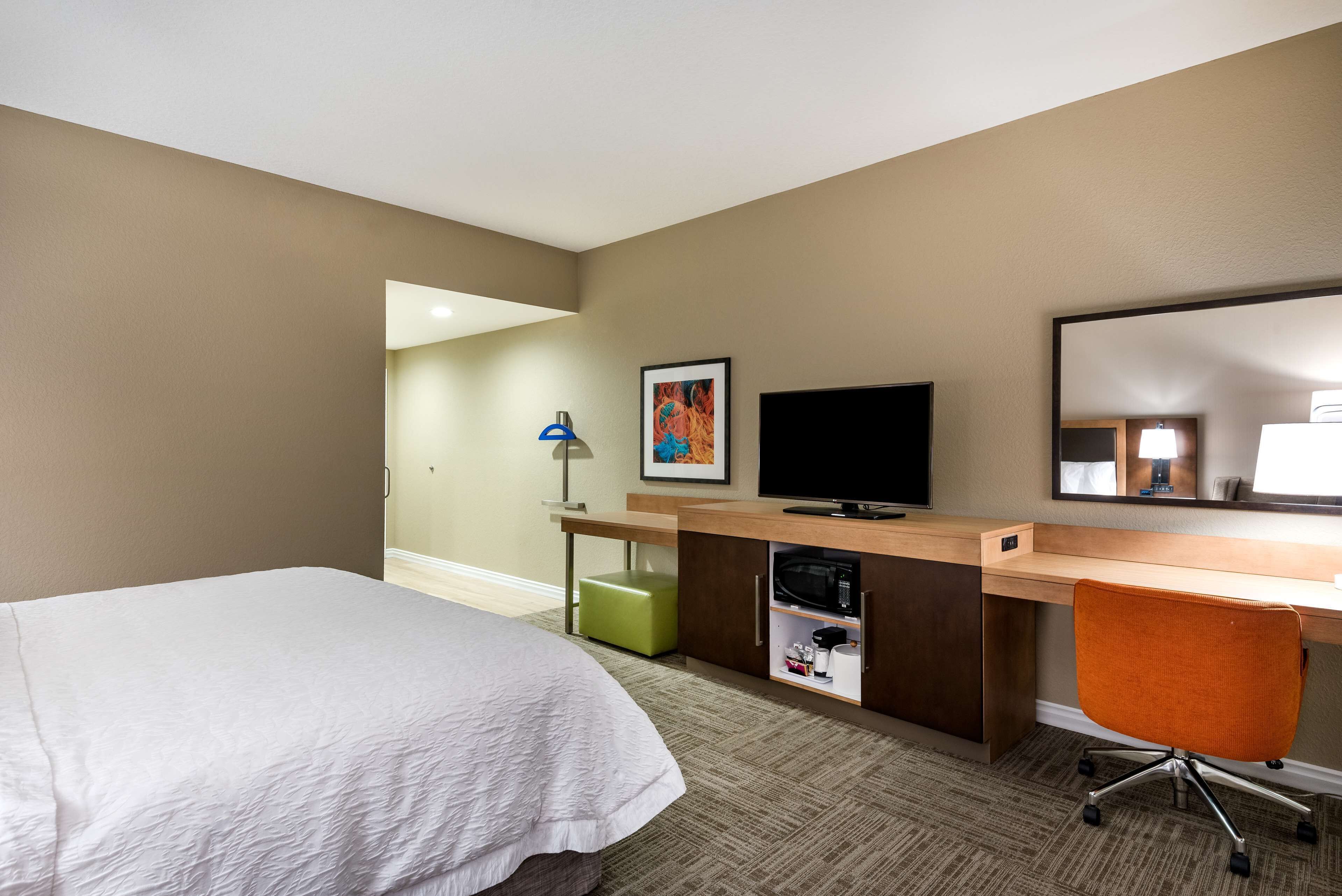 Habitación de hotel con cama, escritorio con televisor, reposapiés verde y espejo.