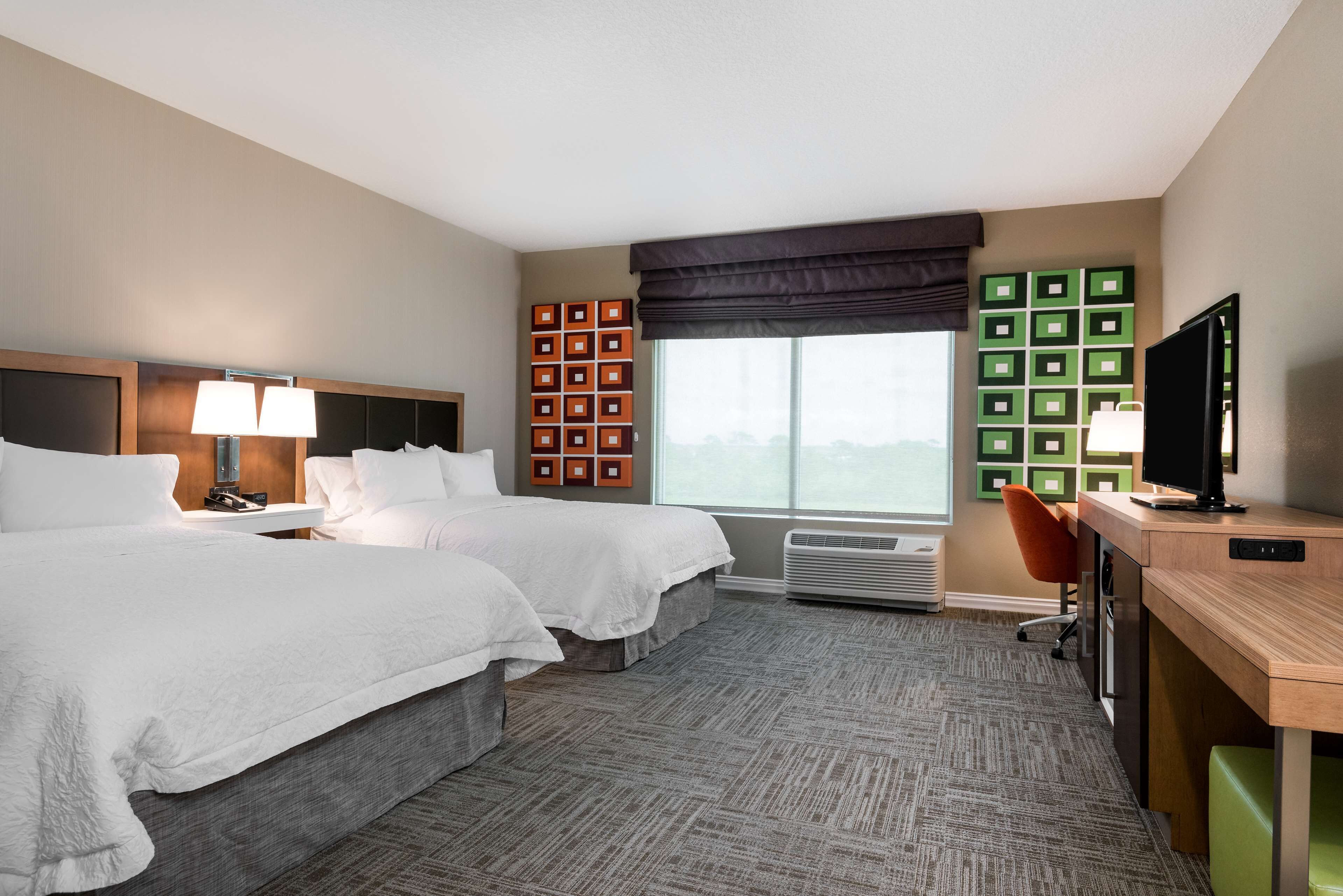 A hotel room with two beds, a desk with a computer, a TV, and a large window with a view.