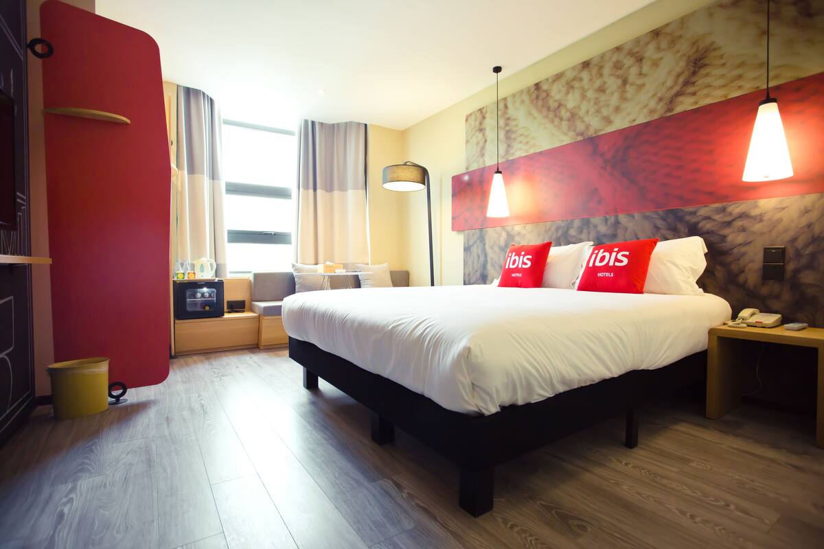 Et hotelværelse med seng, natborde, lampe, garderobe og et vindue med gardiner.