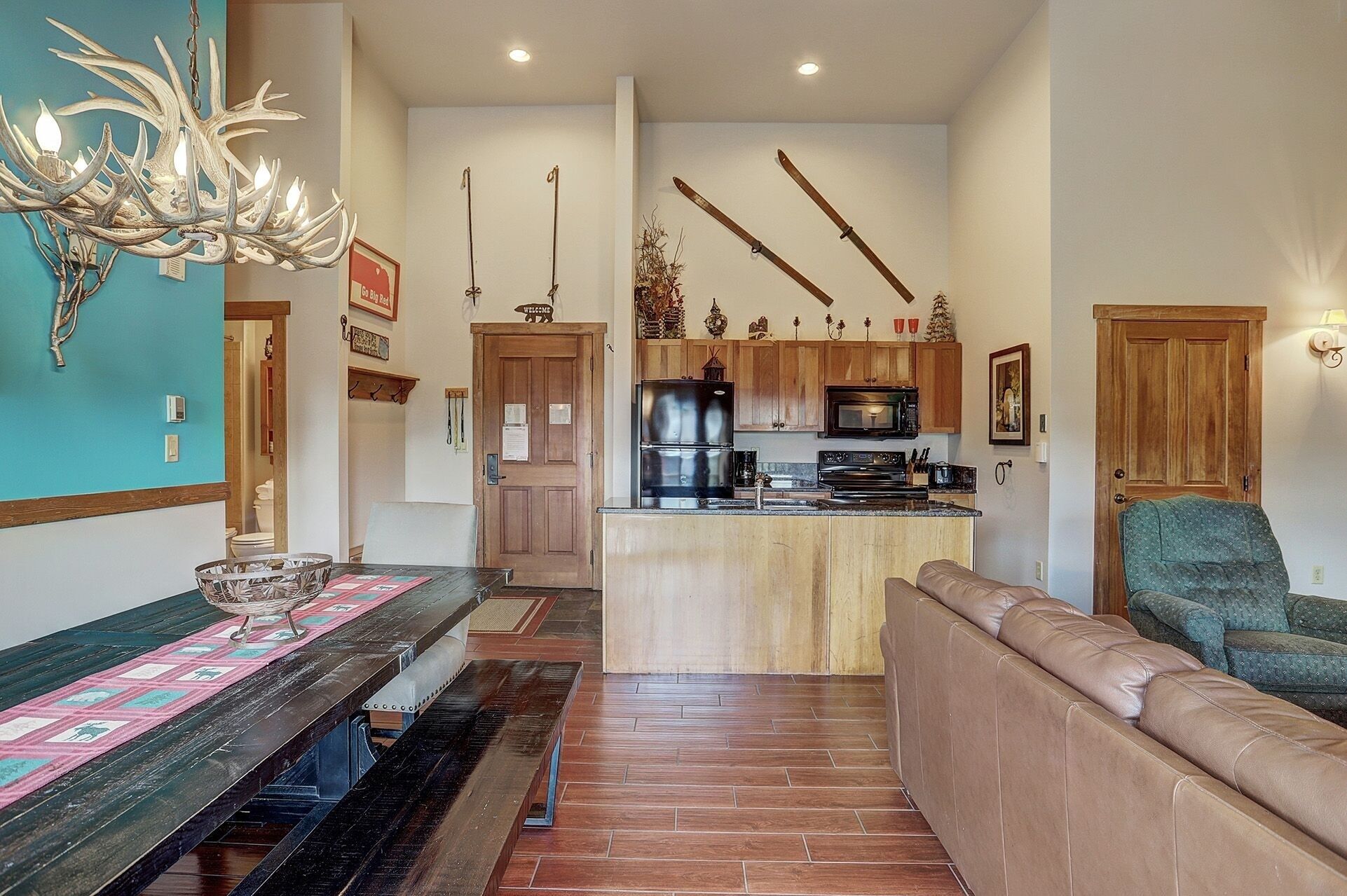 A spacious living area with a wooden bench, a dining table with a placemat, a kitchen with wooden cabinets, and a ski-themed wall decoration.