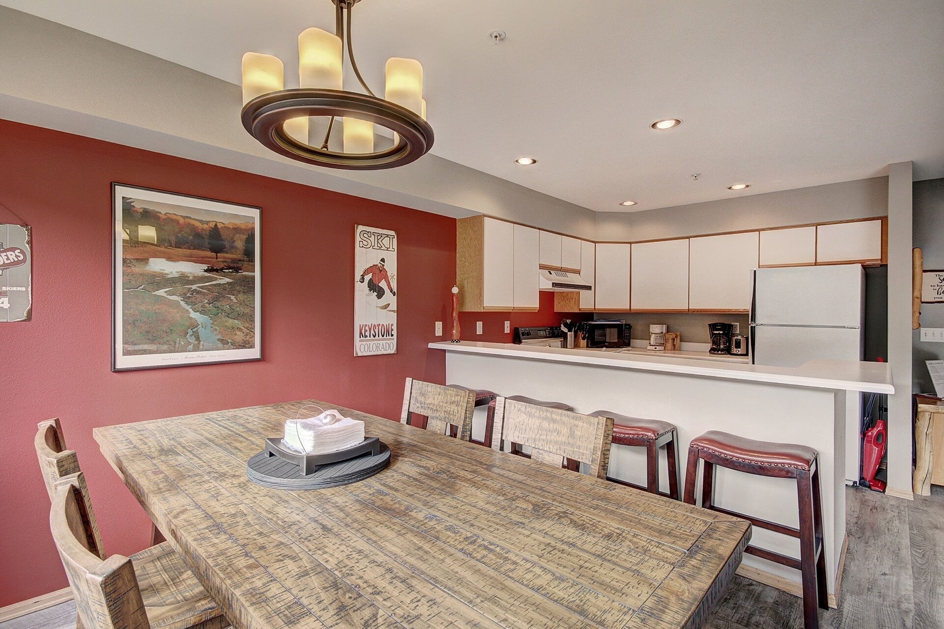 A dining area with a wooden table and chairs, a kitchen with white cabinets, and a mounted ski poster.