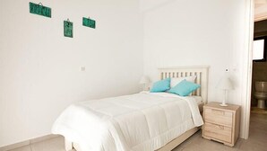 A bedroom with a white bed, two blue pillows, a wooden nightstand, and a white lamp. - Daphne-Morfi Village (Apokoronas)