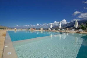 En klarblå pool med solstolar och parasoller, omgiven av berg.