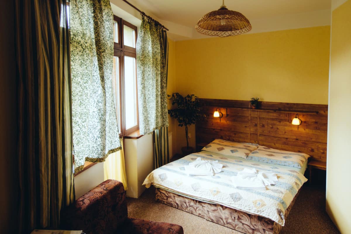 A bedroom with a wooden headboard, a bed with a patterned bedspread, a bedside table with a lamp, a window with curtains, and a small bench.