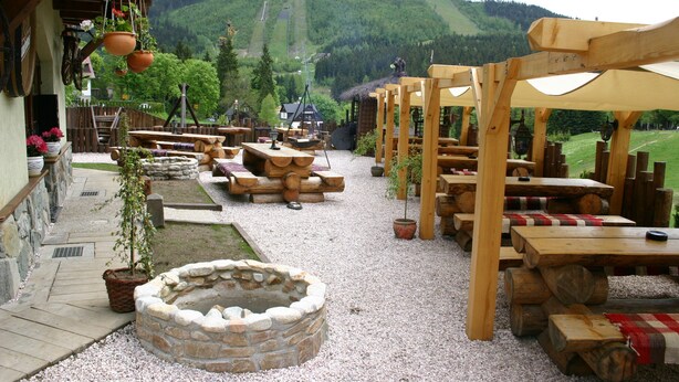 An outdoor seating area with wooden tables and benches, surrounded by potted plants and a stone fire pit. - Pension Rotunda (Harrachov)