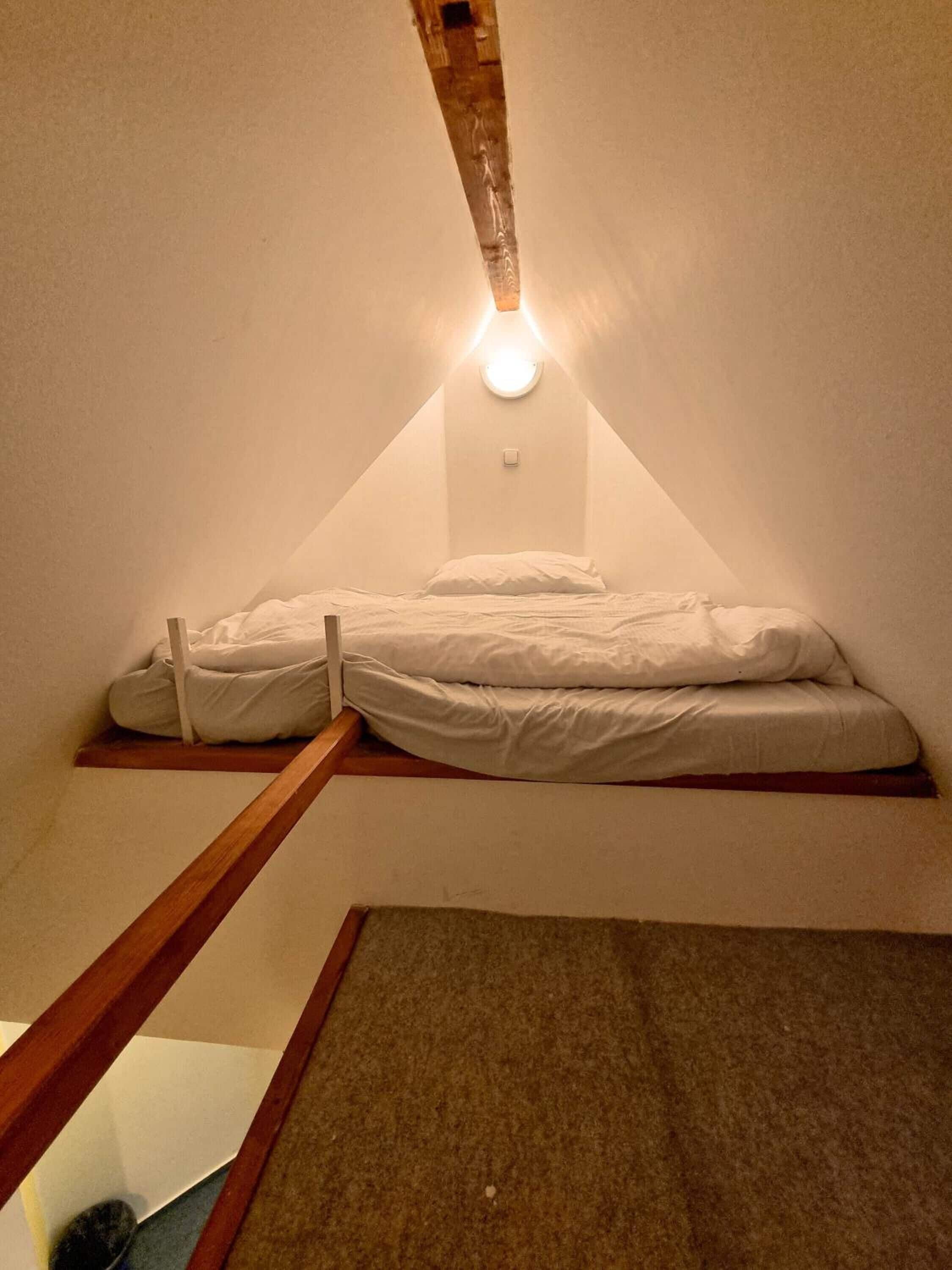 Ein kleines, dreieckiges Schlafzimmer im Dachgeschoss mit einem Bett, einer Holzleiter und einer Leuchte in der geneigten Decke.