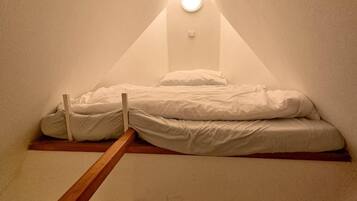 Um quarto de sótão pequeno e triangular, com uma cama de solteiro, uma escada de madeira e um ponto de luz no teto inclinado.