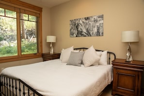 A bedroom with a bed, two bedside tables with lamps, a window with a view of greenery, and a wall-mounted abstract painting.