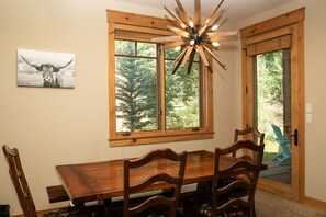 A dining area with a wooden table and chairs, a ceiling light fixture, and a window with a view of trees.