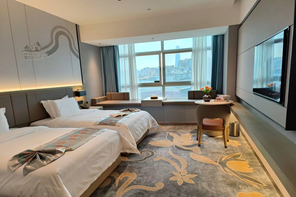 A hotel room with two beds, a seating area, a desk, and a large window with a city view.
