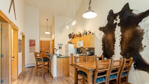 A rustic lodge interior with a wooden dining table, chairs, a kitchen area with wooden cabinets, and a large cowhide wall hanging. - 2674 Tenderfoot Lodge 2 Bedroom Condo by RedAwning (Keystone)