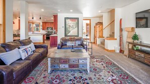 A spacious living room with a leather sofa, a coffee table, and a colorful rug. A kitchen is visible in the background, and there is a staircase leading to another level.