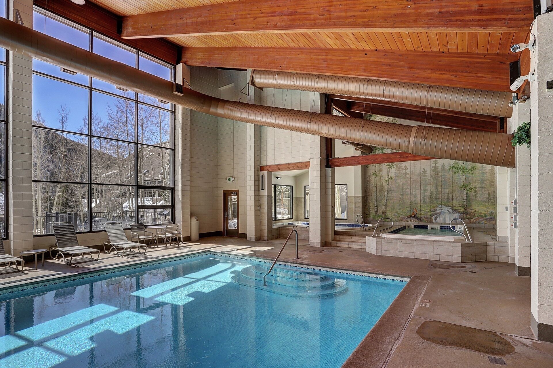 A large indoor swimming pool with a wooden ceiling, large windows, and a view of trees outside.