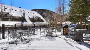 A snow-covered outdoor seating area with metal chairs and tables, a barbecue grill, and a building with a sloped roof in the background. - 2062 The Pines 2 Bedroom Condo by RedAwning (Keystone)