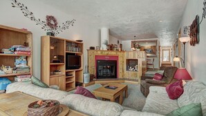 A cozy living room with a fireplace, wooden shelves, a sofa, and a dining area. - 2062 The Pines 2 Bedroom Condo by RedAwning (Keystone)