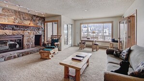 A cozy living room with a stone fireplace, a wooden coffee table, a sofa, and a view of a snowy landscape through large windows. - 221 Cinnamon Ridge I 2 Bedroom Condo by RedAwning (Keystone)