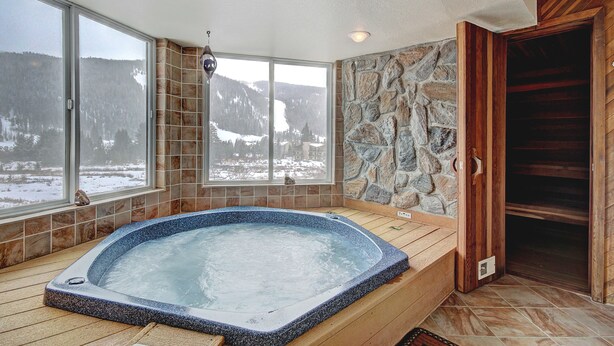 A hot tub with a stone wall and wooden floor, large windows with a snowy mountain view, and a wooden door. - 221 Cinnamon Ridge I 2 Bedroom Condo by RedAwning (Keystone)