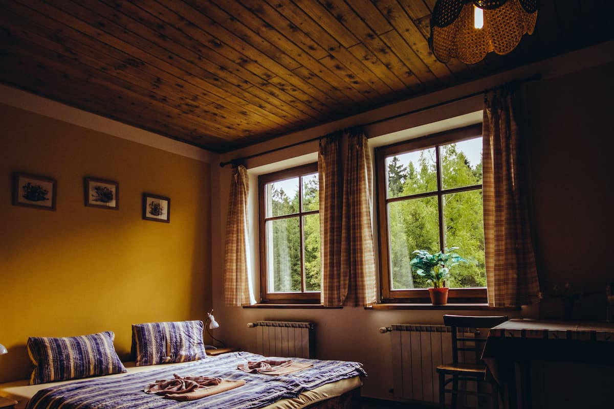 A cozy bedroom with a wooden ceiling, a large bed with patterned bedding, a desk, and a window with curtains.