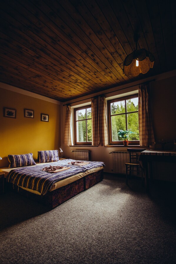 A cozy bedroom with a wooden ceiling, a large bed with patterned bedding, a desk, and a window with curtains.