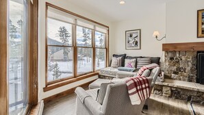 A cozy living room with a stone fireplace, a large window with a view of a snowy landscape, and a comfortable seating area with a plaid throw blanket. - 1655 Quicksilver 3 Bedroom Condo (Keystone)