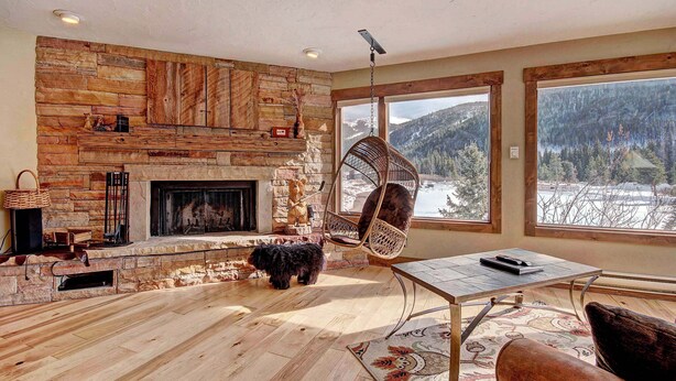 A cozy living room with a stone fireplace, a hanging chair, and large windows offering a view of a snowy landscape.